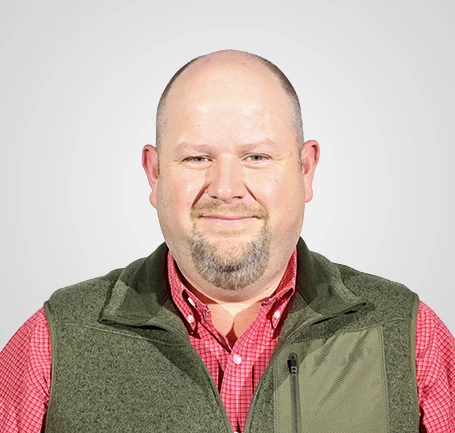 A man with a shaved head and short beard, wearing a red checkered shirt and a green vest, smiling at the camera against a plain light gray background.