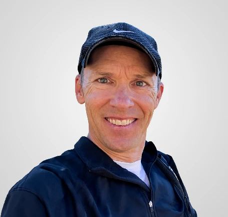 Smiling man wearing a black jacket and a black cap, standing against a plain light gray background.