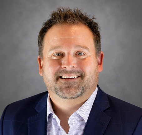 Smiling man with short, tousled brown hair and a beard, wearing a dark blue suit jacket over a light checked shirt, posed in front of a plain gray background. He exudes professionalism and confidence—qualities essential for those passionate about water and soil management.