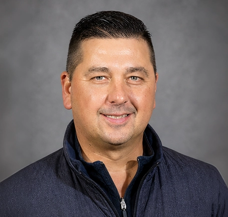 A man with short dark hair wearing a dark zip-up jacket and light blue shirt, smiling in front of a plain grey background, embodying expertise in water and soil management.