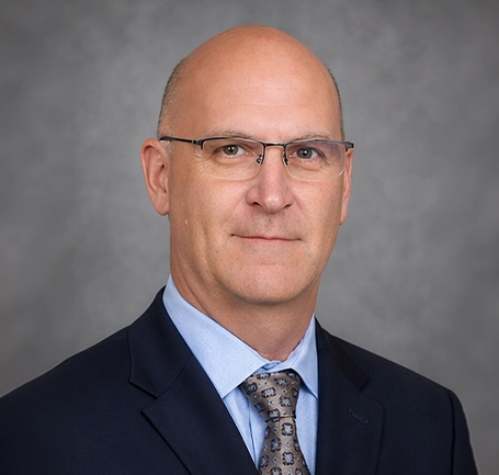 Bald man with glasses wearing a dark suit, light blue shirt, and patterned tie poses in front of a gray background, reflecting his expertise in water and soil management.