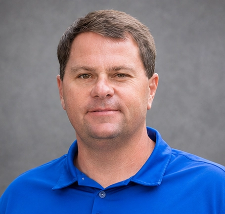 A man with short brown hair wearing a blue polo shirt stands in front of a plain gray background, looking at the camera with a neutral expression, as if ready to discuss topics such as water and soil management.