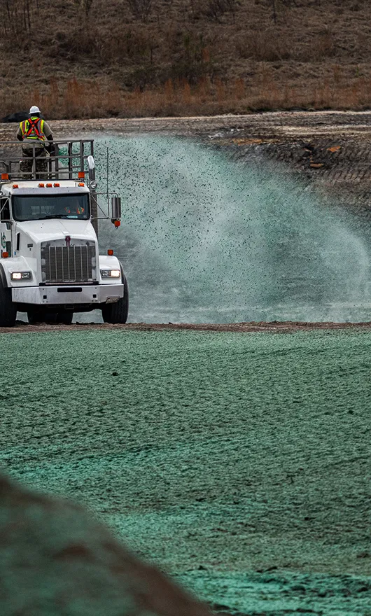 A worker stands on a truck spraying green hydroseed over bare ground, likely for erosion control or re-vegetation, with a dry, brown hillside in the background.