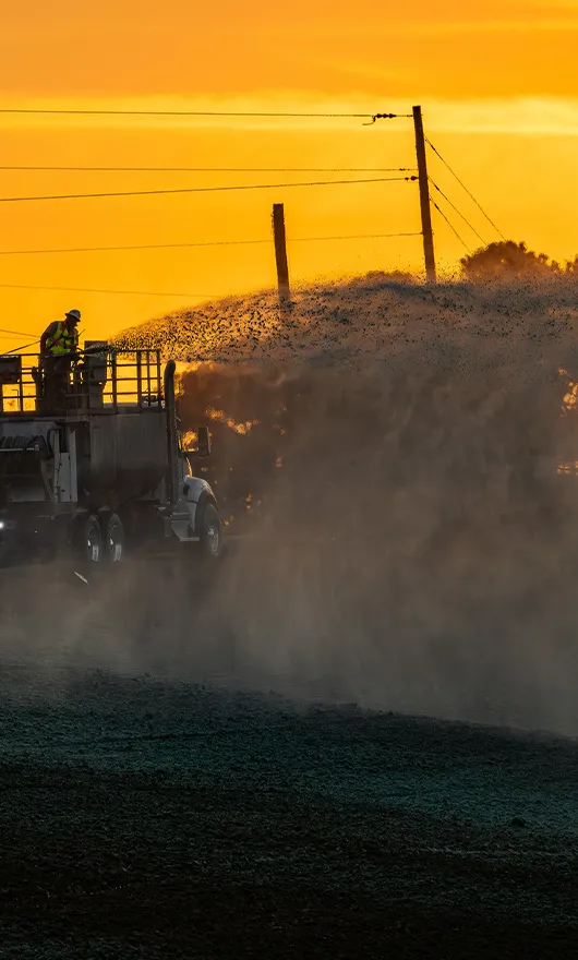 A construction truck sprays water on a dusty road at sunset, with a worker standing on top. Power lines and orange sky are visible in the background.