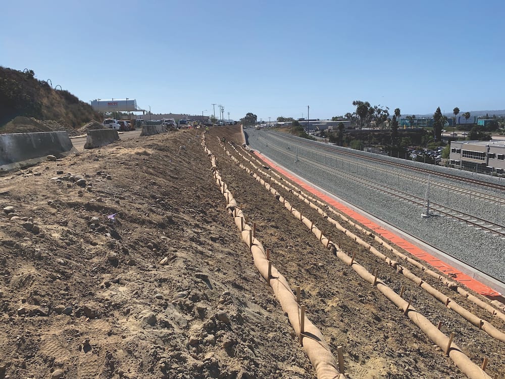 Construction site beside a road with erosion control mats and tubes on a dirt slope. Vehicles and equipment are visible in the background. The sky is clear, and buildings are seen to the right.