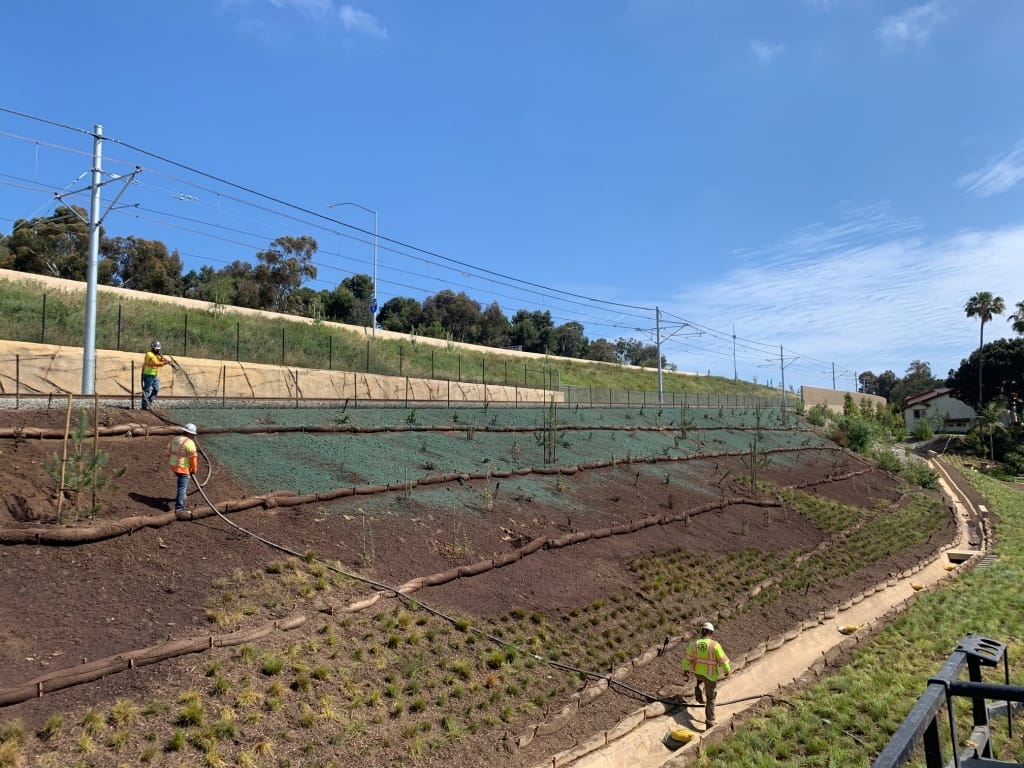 Three workers in safety vests are landscaping a sloped area beside a railway track, planting grass and shrubs under a clear blue sky. Logs and mesh are used for erosion control on the hill.