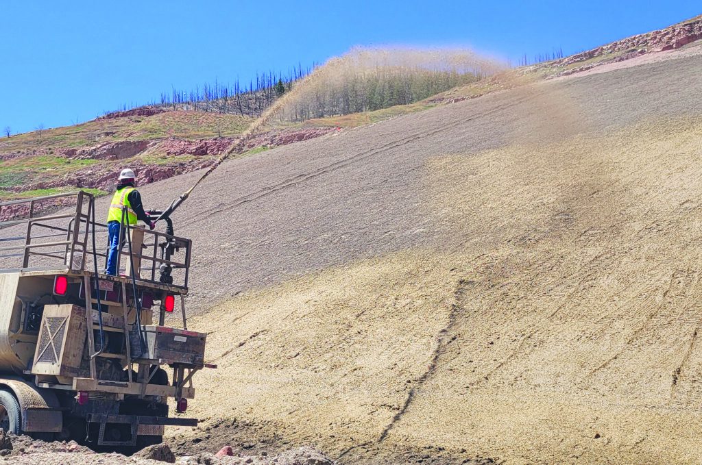 A worker in a safety vest and helmet sprays a hose from a truck onto a steep, barren hillside under a clear blue sky, possibly for erosion control or soil treatment.