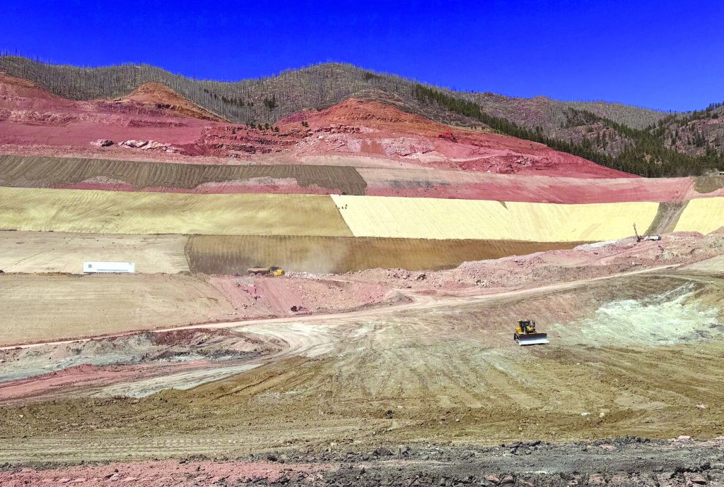 A large open-pit mine with earthmoving machinery working on terraced, multicolored soil layers. Hills and sparse trees are visible in the background under a clear blue sky.