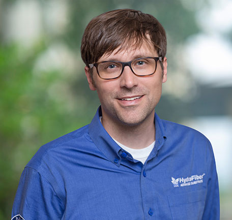 A man with brown hair and glasses smiles, wearing a blue HydraFiber shirt. The background is softly blurred with green and white tones.