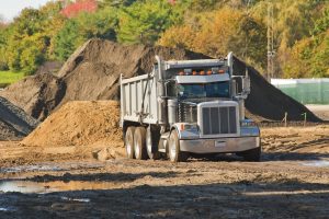 A silver dump truck is parked at a construction site with dirt piles and muddy ground, surrounded by trees with green and autumn-colored leaves in the background.