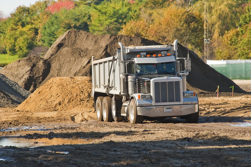 A silver dump truck is parked at a construction site with dirt piles and muddy ground, surrounded by trees with green and autumn-colored leaves in the background.