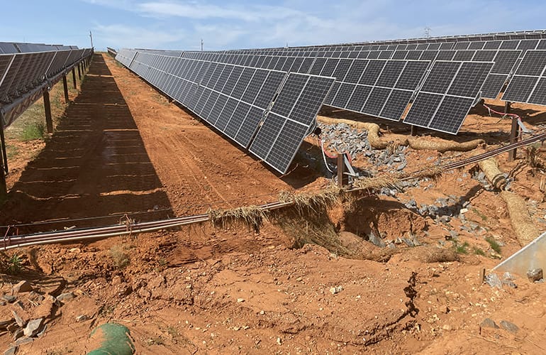 Rows of solar panels on a reddish dirt field; a large eroded ditch cuts through the ground beneath metal pipes, exposing roots and rocks.