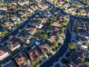 Aerial view of a suburban neighborhood with winding streets, houses with brown roofs, green lawns, palm trees, and cars parked along the roads on a sunny day.