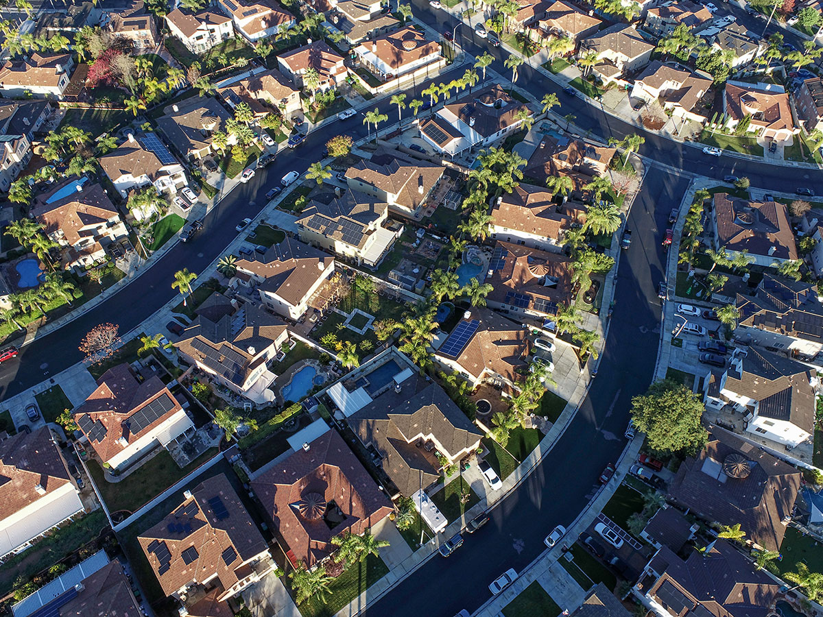 Aerial view of a suburban neighborhood with winding streets, houses with brown roofs, green lawns, palm trees, and cars parked along the roads on a sunny day.