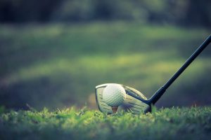 A close-up of a golf ball on a tee with a driver club positioned behind it, ready to hit, on a grassy course with a blurred background.