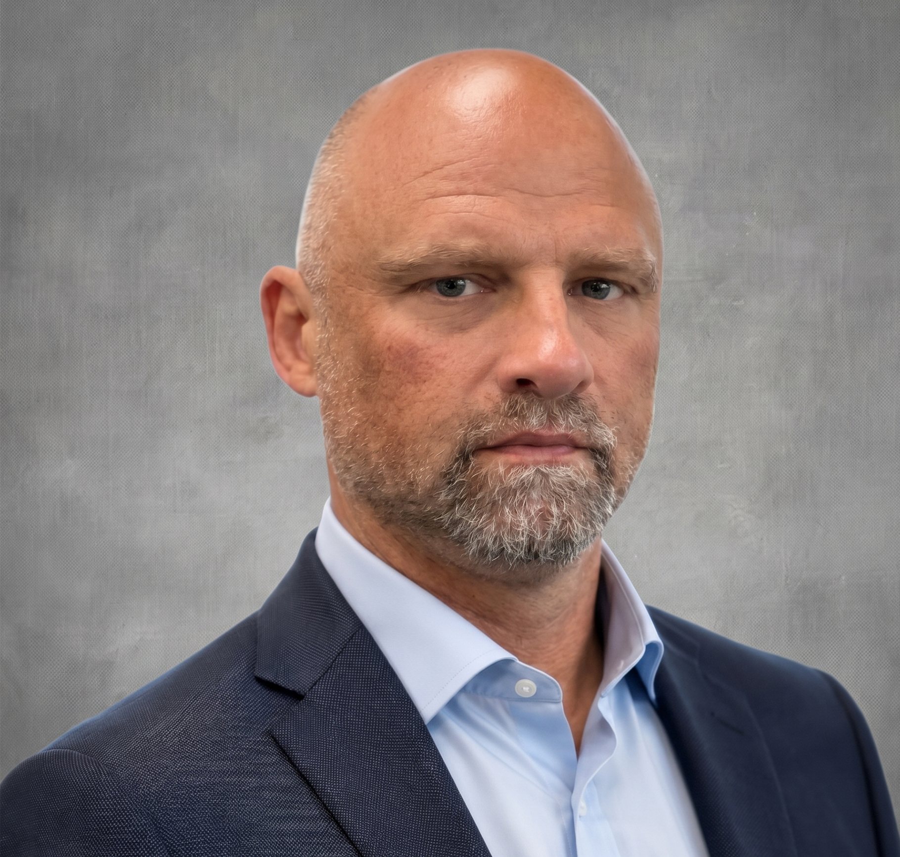 A middle-aged man with a shaved head and short beard, wearing a navy blue suit and light blue shirt, stands in front of a blurred office background, looking serious—reflecting his dedication to water and soil management.