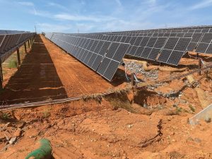 Rows of solar panels on reddish soil, with visible erosion causing a gully and exposed pipes beneath some panels. Clear blue sky overhead.