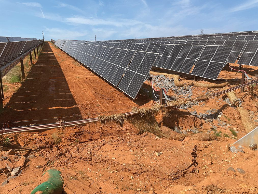 Rows of solar panels on reddish soil, with visible erosion causing a gully and exposed pipes beneath some panels. Clear blue sky overhead.