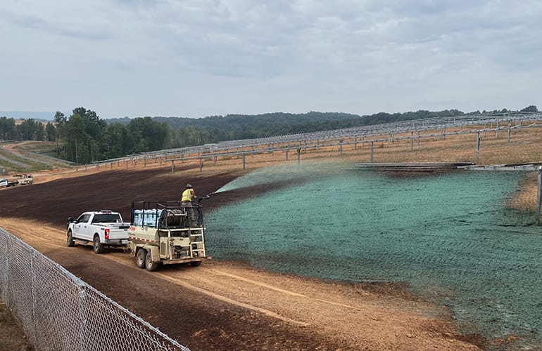 A truck sprays green hydroseed on a dirt slope near a fenced road, with solar panel frames and trees in the background under a cloudy sky.