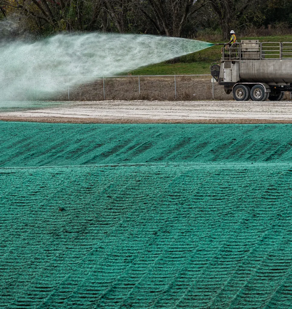 A worker on a truck sprays green hydroseeding material over a large, cleared field near a fence and trees, promoting new grass growth.