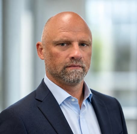 A middle-aged man with a shaved head and short beard, wearing a navy blue suit and light blue shirt, stands in front of a blurred office background, looking serious—reflecting his dedication to water and soil management.