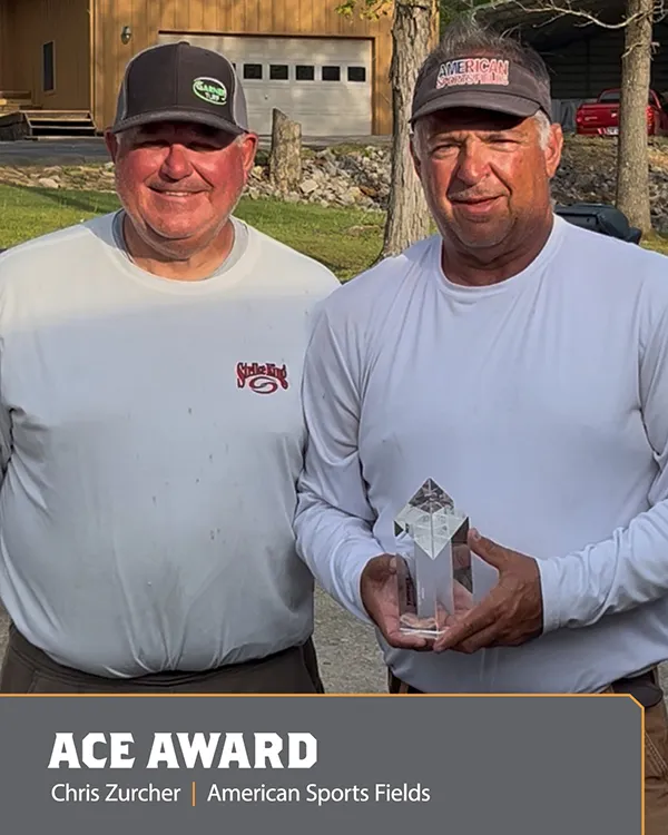 Two men stand outdoors, one holding a crystal award. Both wear light shirts and caps, smiling at the camera. Text reads: ACE AWARD. Chris Zurcher | American Sports Fields..