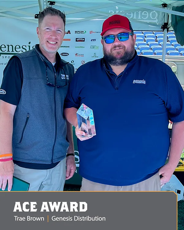 Two men stand together under a tent; one holds a crystal award. Both are smiling and wearing Genesis-branded shirts. A sign behind them displays logos. Caption reads ACE AWARD Trae Brown | Genesis Distribution..