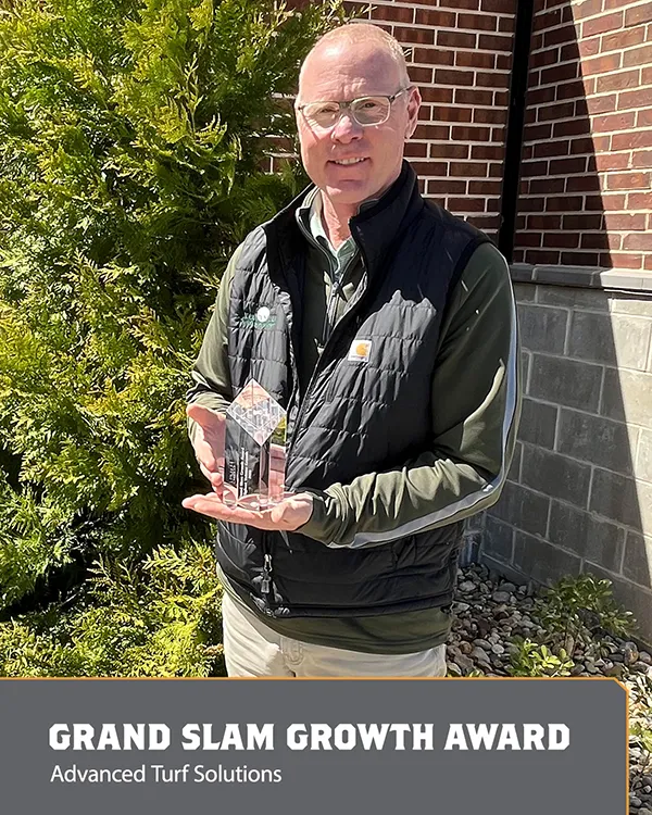 A man standing outside in front of greenery and a brick wall holds a clear glass trophy. A sign at the bottom reads: GRAND SLAM GROWTH AWARD, Advanced Turf Solutions..