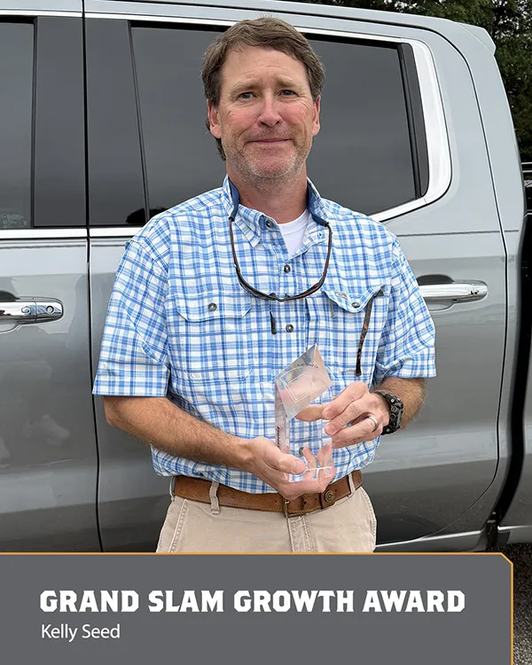 A man in a blue plaid shirt stands in front of a silver vehicle, holding a clear award. Text below reads: GRAND SLAM GROWTH AWARD, Kelly Seed..