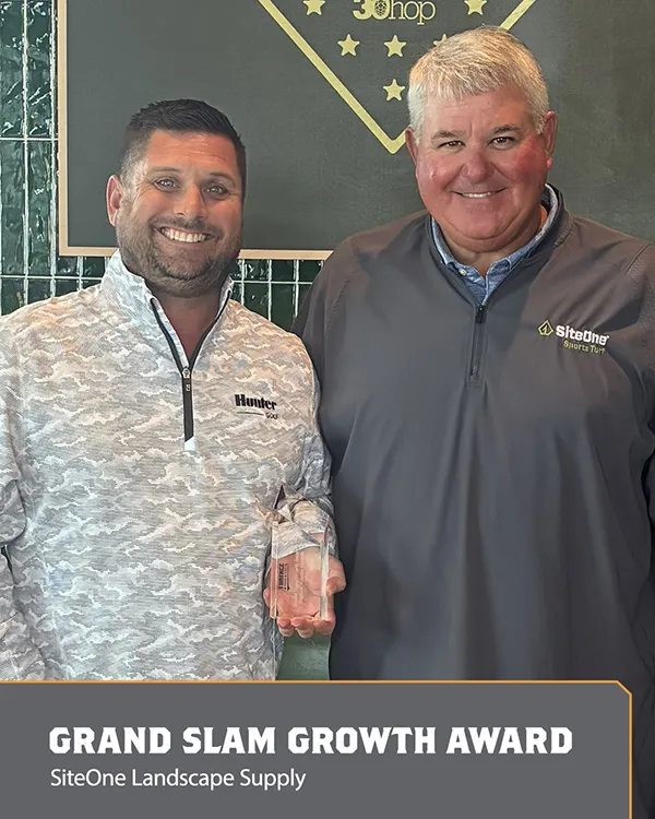 Two smiling men stand together, one holding a glass award. Both wear gray jackets with company logos. A sign below reads Grand Slam Growth Award, SiteOne Landscape Supply..