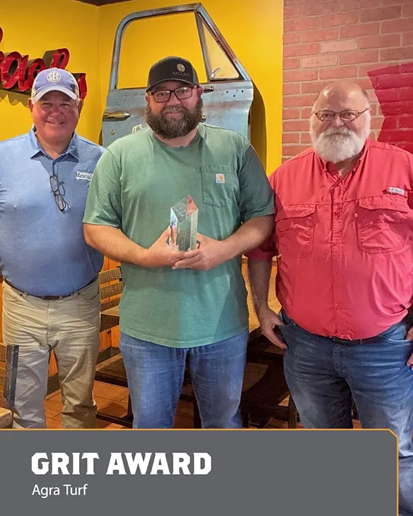 Three men stand indoors; the center man holds a glass award. Text at the bottom reads, GRIT AWARD Agra Turf. A blue truck door and yellow and brick walls are in the background.