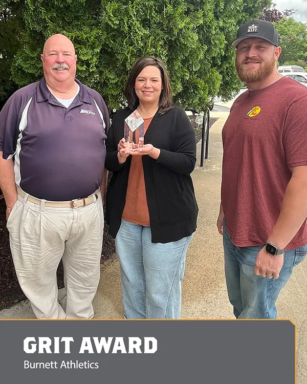 Three people stand outdoors, smiling, as the woman in the middle holds a clear trophy. A sign at the bottom reads GRIT AWARD - Burnett Athletics. Green trees are in the background.