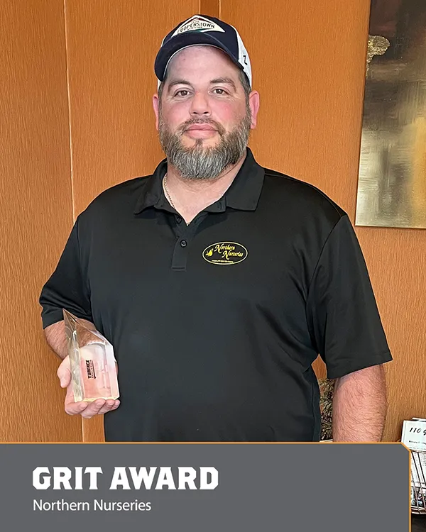 A bearded man in a black polo and cap stands indoors, holding a clear trophy. Text at the bottom reads, GRIT AWARD Northern Nurseries..