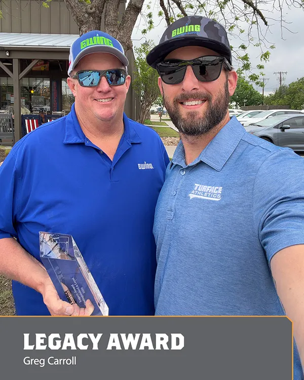 Two men in blue shirts and sunglasses pose outdoors. One holds a clear award. A graphic at the bottom reads LEGACY AWARD and Greg Carroll..
