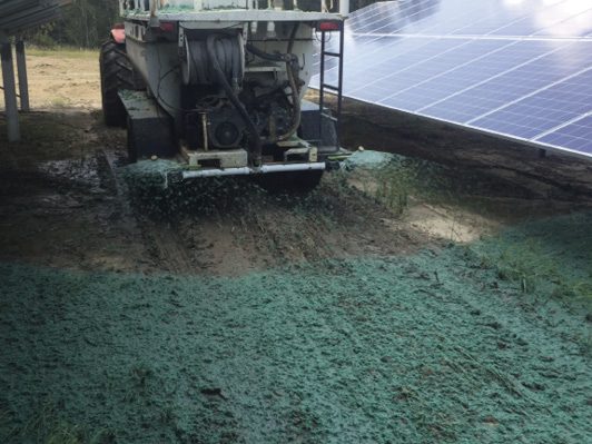 A truck sprays green hydroseed onto the ground beneath solar panels, covering soil and grass to promote new vegetation growth.