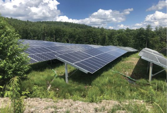 Rows of solar panels are installed in a grassy, open area with trees and a forested hill in the background under a partly cloudy sky.