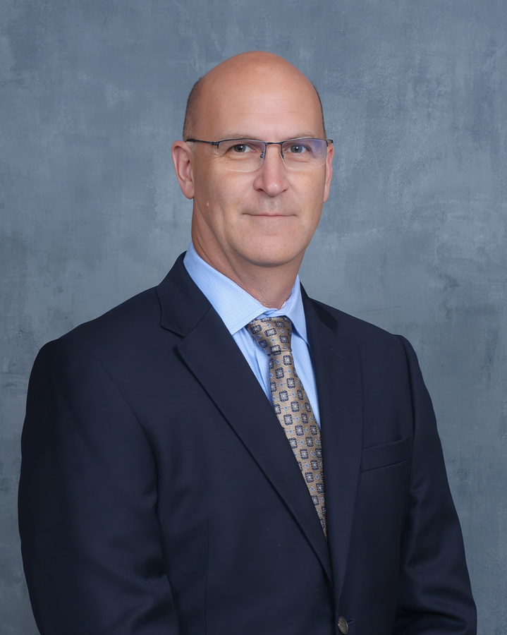 A man wearing glasses, a dark suit, blue shirt, and patterned tie stands in front of a gray textured background, facing forward with a neutral expression.