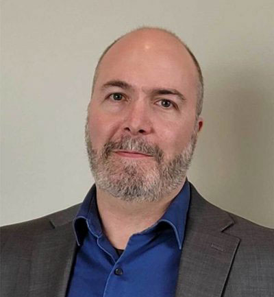 A man with a shaved head and short gray beard is wearing a blue shirt and gray blazer, standing in front of a plain, light-colored background.