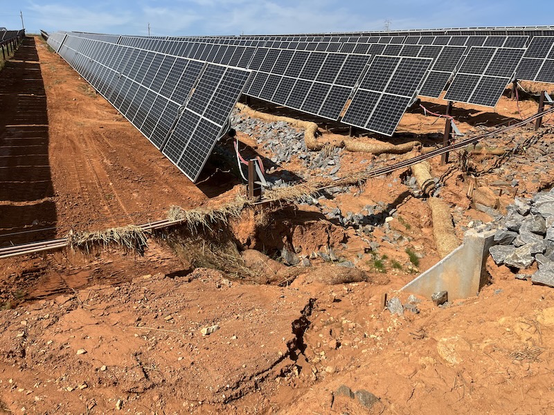 A row of solar panels stands above eroded red soil, with exposed pipes and rocks visible where the ground has washed away, creating a noticeable gap and damage.