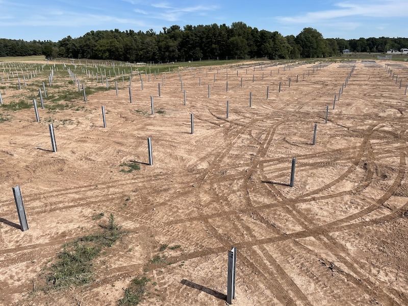 A dirt field with evenly spaced metal posts sticking out of the ground, tire tracks visible, and trees in the background under a partly cloudy sky.