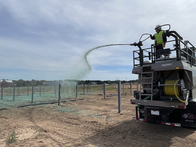 A worker in a safety vest operates a hydroseeding truck, spraying green mulch onto a large, sandy construction site with metal fence posts under a partly cloudy sky.