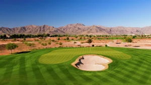 A green golf course with a sand bunker in the foreground, surrounded by mountains and clear blue sky in the background.