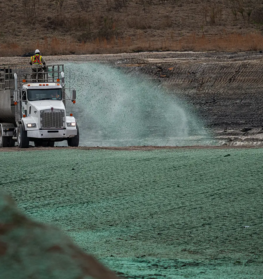 A truck sprays green hydroseed onto bare soil while a worker in a safety vest and helmet stands on top.
