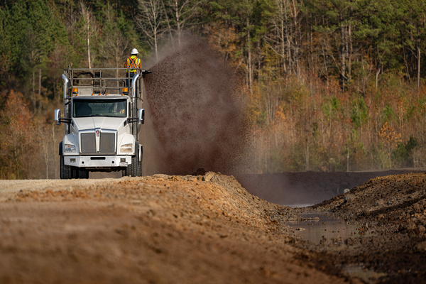 A construction truck dumps a load of dirt on a road while a worker in a safety vest stands on top. Trees with autumn foliage fill the background.