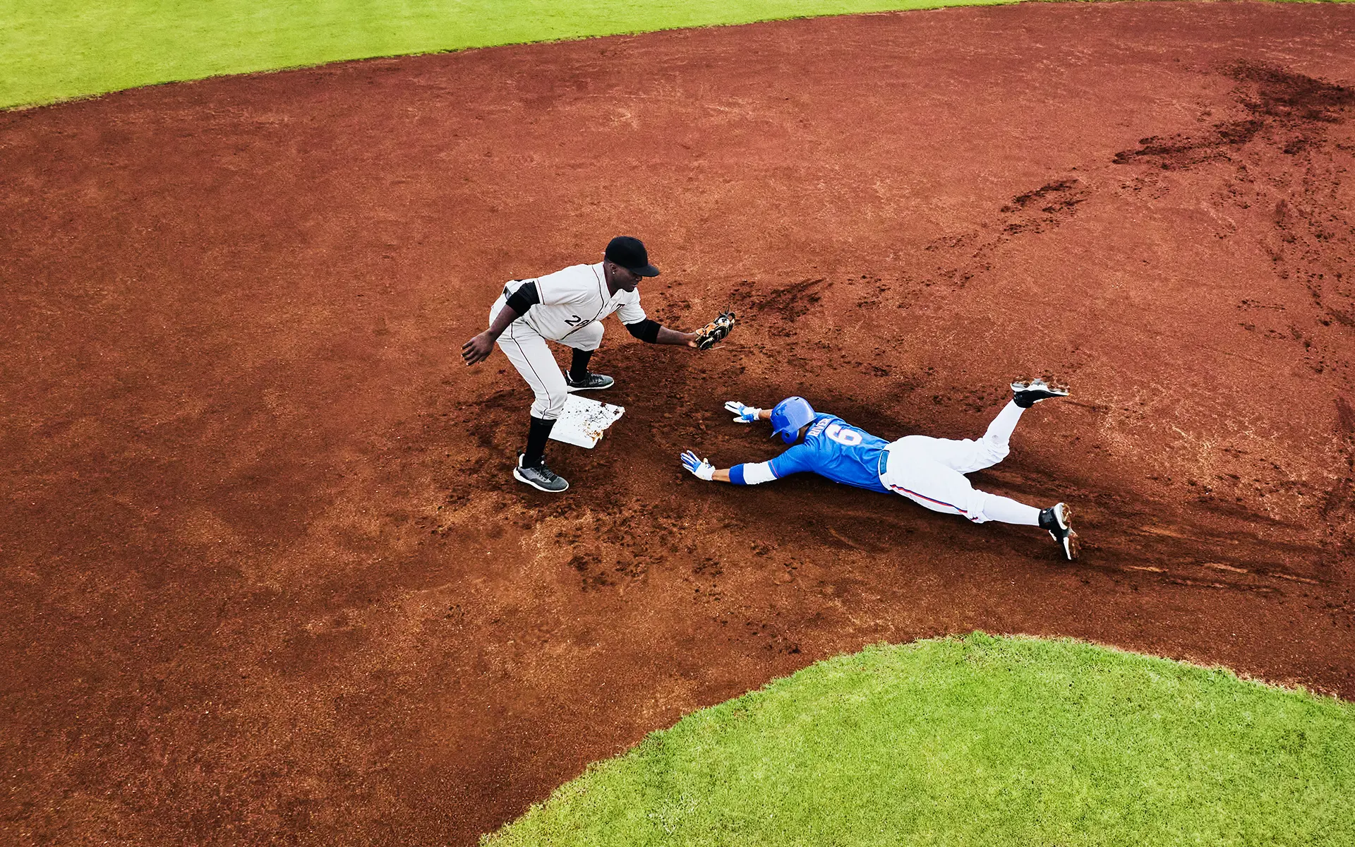 A baseball player in blue slides headfirst into a base as a player in white attempts to tag him out on a dirt infield, with grass visible at the edges.