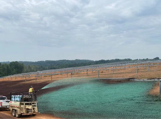 A worker sprays green hydroseed on bare soil near metal framework structures under a cloudy sky, with trucks parked nearby and trees in the background.