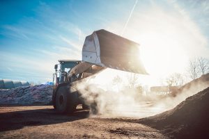 A large front loader lifts dirt at a construction site on a sunny day, with dust rising around its wheels and the sun shining brightly in the background.