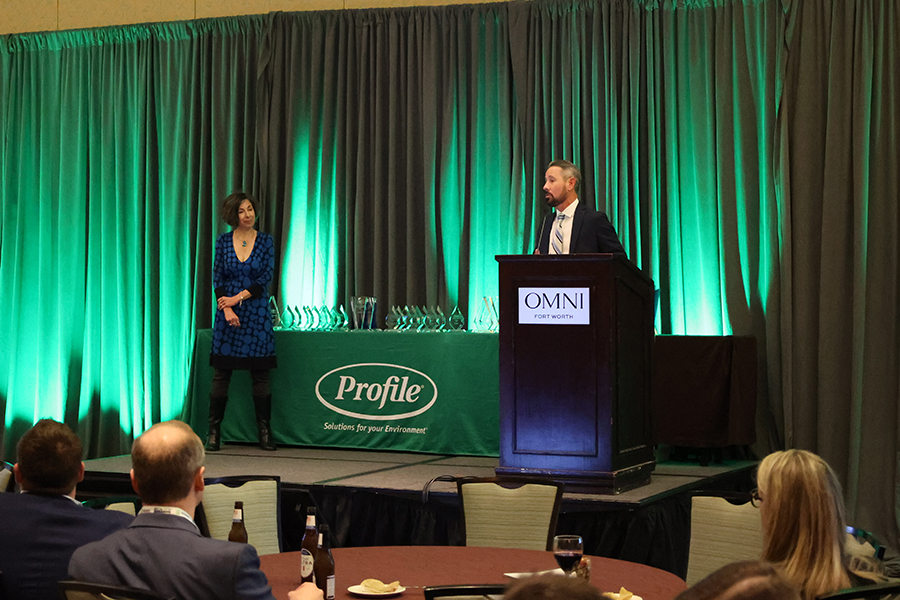 Man speaking at a podium labeled Omni as a woman stands nearby on stage; numerous glass awards are displayed on a table with a green Profile banner; audience seated in the foreground.