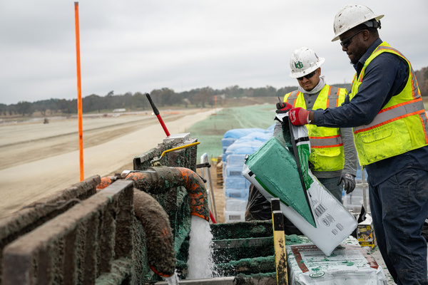 Two construction workers in safety vests and helmets pour material from a bag into machinery at a work site, with equipment and bags in the background. The scene appears overcast and industrial.