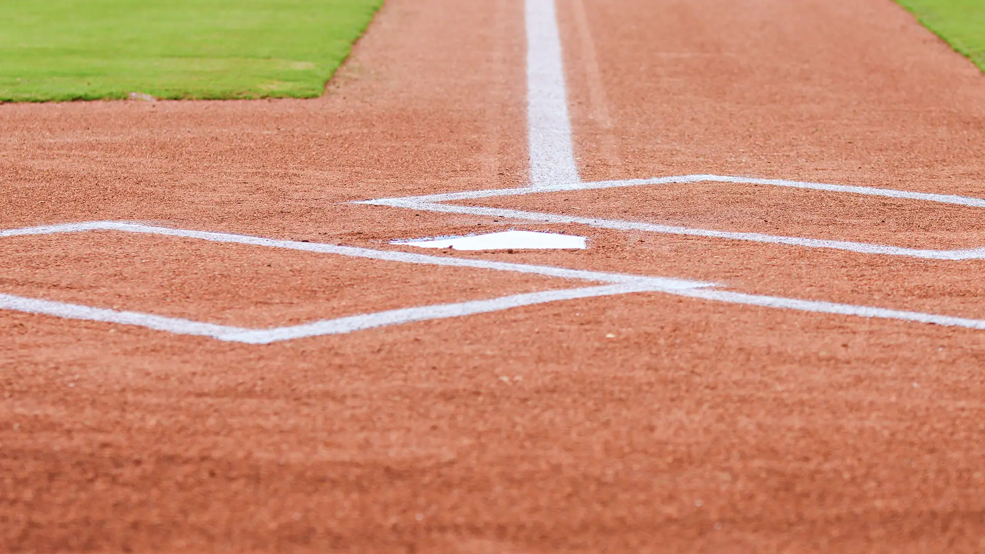 Close-up of a baseball fields home plate area with white chalk lines marking the batters box, part of the infield dirt, and green grass visible in the upper left corner.